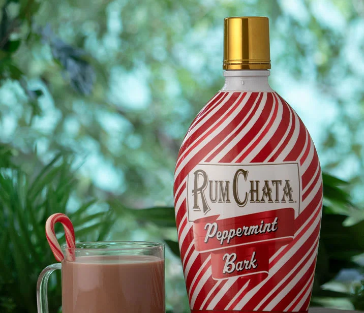 A glass cup of peppermint bark hot chocolate with a candy cane and a bottle Rumchata peppermint bark